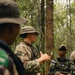 The image shows military personnel in a forested environment, appearing to conduct a briefing or instruction session. A central figure wearing MultiCam/OCP camouflage with a US flag patch and boonie hat is gesturing with their hands, apparently explaining something to others. The setting and uniforms suggest a joint training exercise involving at least two different military forces.