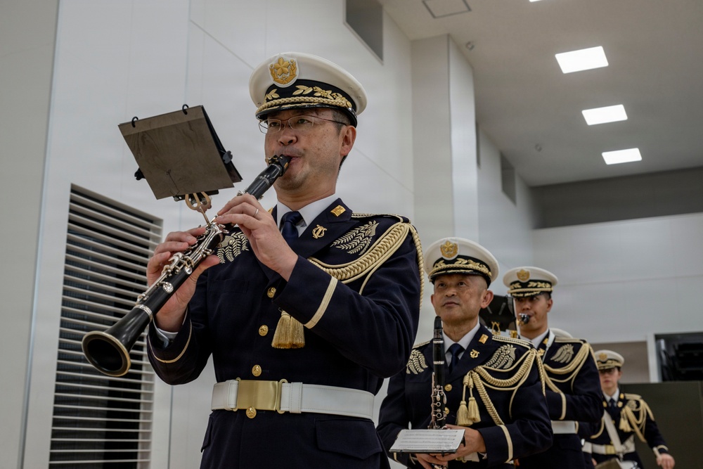 A Japanese military or police band is performing indoors, with the foreground musician playing a clarinet while reading from a music stand. The musicians are dressed in formal dark navy ceremonial uniforms with gold epaulettes, braiding, and white peaked caps. At least three additional band members are visible in the background, also in identical uniforms.