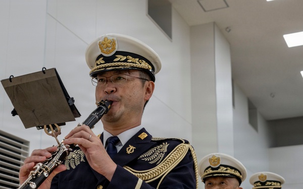 A Japanese military or police band is performing indoors, with the foreground musician playing a clarinet while reading from a music stand. The musicians are dressed in formal dark navy ceremonial uniforms with gold epaulettes, braiding, and white peaked caps. At least three additional band members are visible in the background, also in identical uniforms.