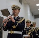 A Japanese military or police band is performing indoors, with the foreground musician playing a clarinet while reading from a music stand. The musicians are dressed in formal dark navy ceremonial uniforms with gold epaulettes, braiding, and white peaked caps. At least three additional band members are visible in the background, also in identical uniforms.