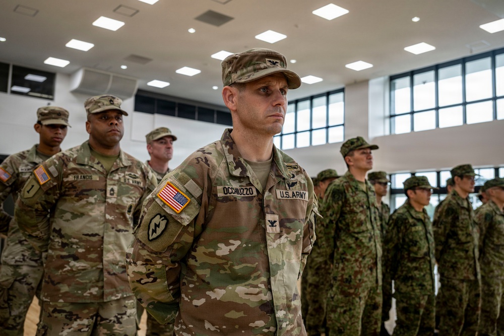 A formal military formation or ceremony is taking place indoors, featuring U.S. Army personnel alongside soldiers in a different camouflage pattern, likely allied or partner nation forces. The central figure, identified by name tape as 'OCCHIUZZO' and wearing a Colonel's eagle insignia, stands at attention in U.S. Army OCP (Operational Camouflage Pattern) uniform. The setting appears to be a modern military facility or gymnasium.
