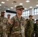 A formal military formation or ceremony is taking place indoors, featuring U.S. Army personnel alongside soldiers in a different camouflage pattern, likely allied or partner nation forces. The central figure, identified by name tape as 'OCCHIUZZO' and wearing a Colonel's eagle insignia, stands at attention in U.S. Army OCP (Operational Camouflage Pattern) uniform. The setting appears to be a modern military facility or gymnasium.
