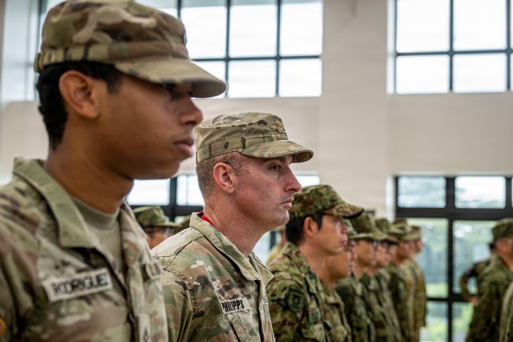 The image shows a formation of military personnel standing at attention inside a large building with tall windows. Two U.S. Army soldiers in OCP (Operational Camouflage Pattern) uniforms are prominently visible in the foreground, with name tapes reading 'RODRIGUE[Z]' and 'PHILIPPS'. Behind them, a row of soldiers wearing a different camouflage pattern — consistent with Japanese Ground Self-Defense Force uniforms — suggests this is a joint military exercise or ceremony.
