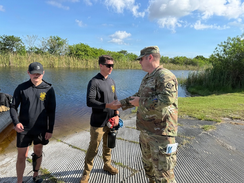 A U.S. Army soldier in full OCP camouflage uniform is engaged in a handshake or exchange with a civilian wearing a black long-sleeve shirt with a yellow logo, while a second civilian in matching black attire stands nearby. The group is standing on a metal grated dock or boat ramp adjacent to a body of water in what appears to be a wetland or marsh environment. The setting suggests a field coordination meeting or briefing near a waterway.