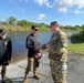 A U.S. Army soldier in full OCP camouflage uniform is engaged in a handshake or exchange with a civilian wearing a black long-sleeve shirt with a yellow logo, while a second civilian in matching black attire stands nearby. The group is standing on a metal grated dock or boat ramp adjacent to a body of water in what appears to be a wetland or marsh environment. The setting suggests a field coordination meeting or briefing near a waterway.