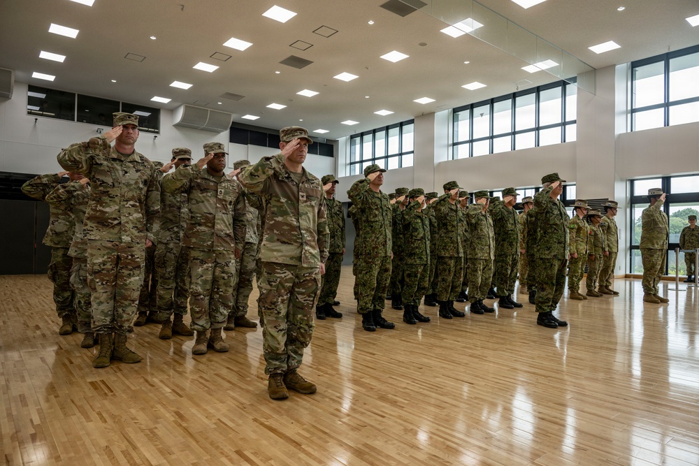 A formal military ceremony is taking place inside a large indoor gymnasium or assembly hall with hardwood floors. Two distinct groups of soldiers in different camouflage uniforms are standing in formation and rendering salutes simultaneously. The left group appears to wear US Army OCP (Operational Camouflage Pattern) uniforms, while the right group wears a different camouflage pattern consistent with Japan Ground Self-Defense Force uniforms, suggesting a joint military exercise or ceremony.