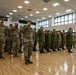 A formal military ceremony is taking place inside a large indoor gymnasium or assembly hall with hardwood floors. Two distinct groups of soldiers in different camouflage uniforms are standing in formation and rendering salutes simultaneously. The left group appears to wear US Army OCP (Operational Camouflage Pattern) uniforms, while the right group wears a different camouflage pattern consistent with Japan Ground Self-Defense Force uniforms, suggesting a joint military exercise or ceremony.