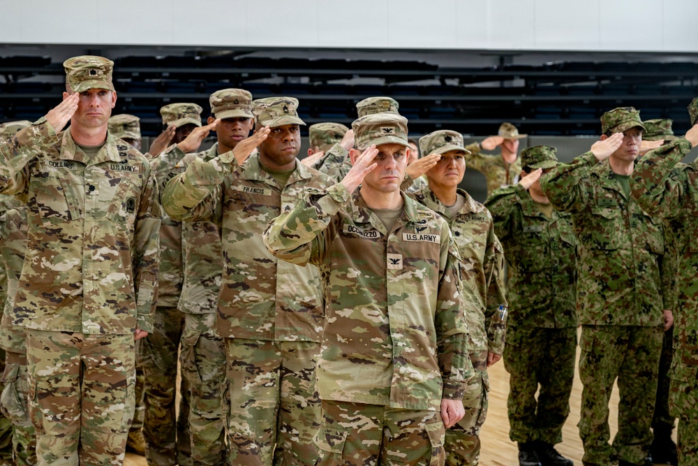 A formal military ceremony showing U.S. Army soldiers and what appear to be Japanese Self-Defense Force personnel rendering salutes simultaneously. The soldiers are standing in organized rows inside what appears to be a gymnasium or indoor facility. Visible name tapes and rank insignia identify several U.S. Army personnel, suggesting a joint military event.