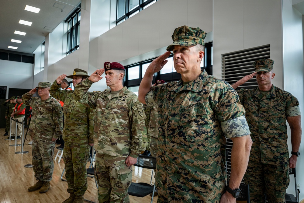A group of military personnel from multiple branches and nations are standing at salute during a formal ceremony held indoors. The foreground features a U.S. Marine Corps officer with two-star general rank insignia, flanked by U.S. Army personnel and other Marines. The setting appears to be a modern military facility or conference center.