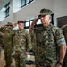A group of military personnel from multiple branches and nations are standing at salute during a formal ceremony held indoors. The foreground features a U.S. Marine Corps officer with two-star general rank insignia, flanked by U.S. Army personnel and other Marines. The setting appears to be a modern military facility or conference center.