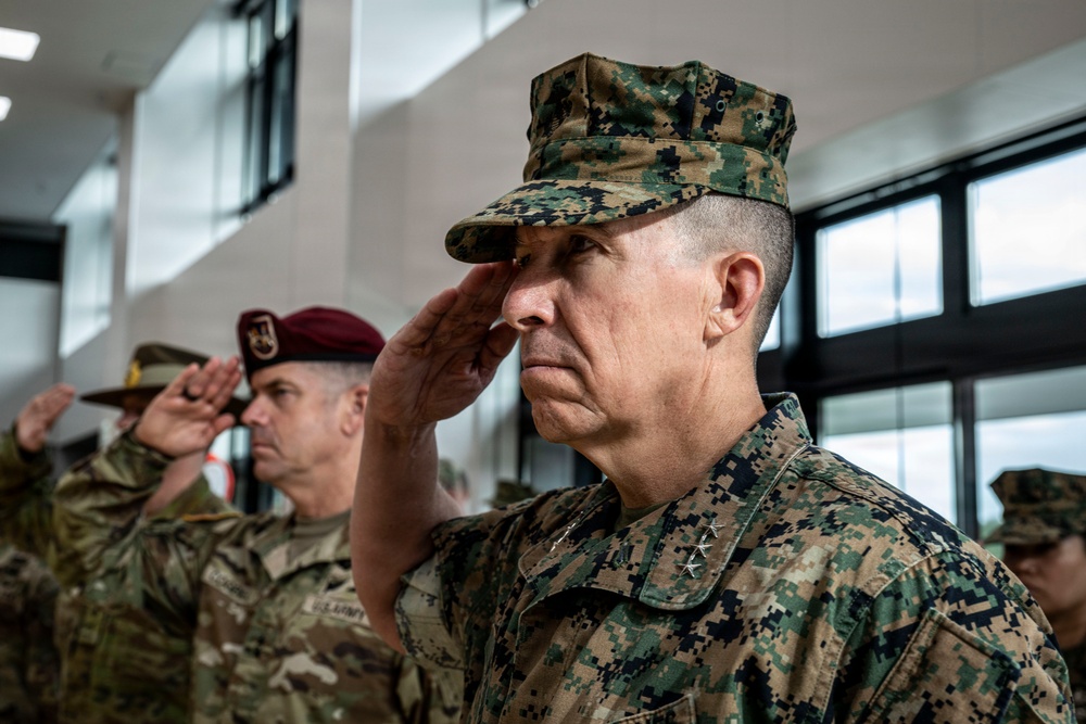 Multiple military personnel in various uniforms are rendering a salute during what appears to be a formal ceremony or official event. The foreground figure wears United States Marine Corps MARPAT digital camouflage with three-star rank insignia (Lieutenant General). Behind him are Army personnel, including one wearing a maroon beret associated with airborne units.