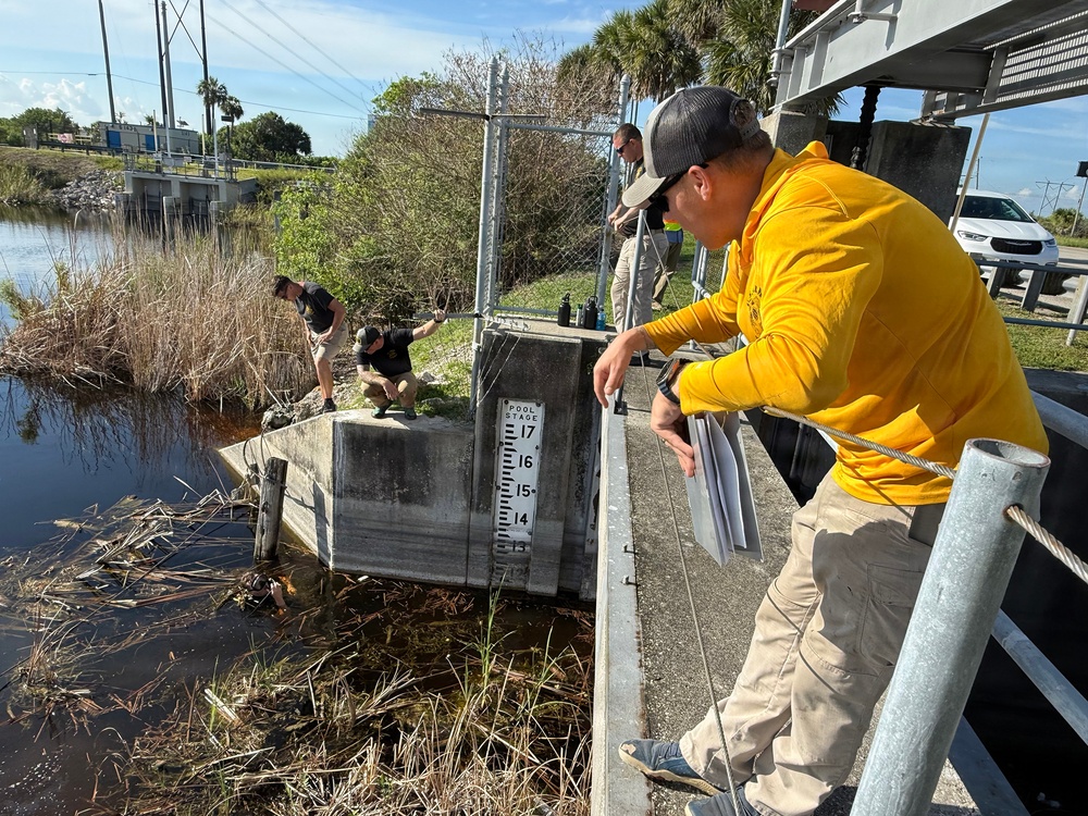 A group of workers or officials are inspecting a water control structure, likely a flood gate or weir, at a waterway in what appears to be Florida based on the palm trees and vegetation. A pool stage gauge is prominently visible on the concrete structure, showing water level markings between 14 and 17. The scene suggests post-storm or routine water management assessment activities.