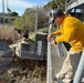 A group of workers or officials are inspecting a water control structure, likely a flood gate or weir, at a waterway in what appears to be Florida based on the palm trees and vegetation. A pool stage gauge is prominently visible on the concrete structure, showing water level markings between 14 and 17. The scene suggests post-storm or routine water management assessment activities.