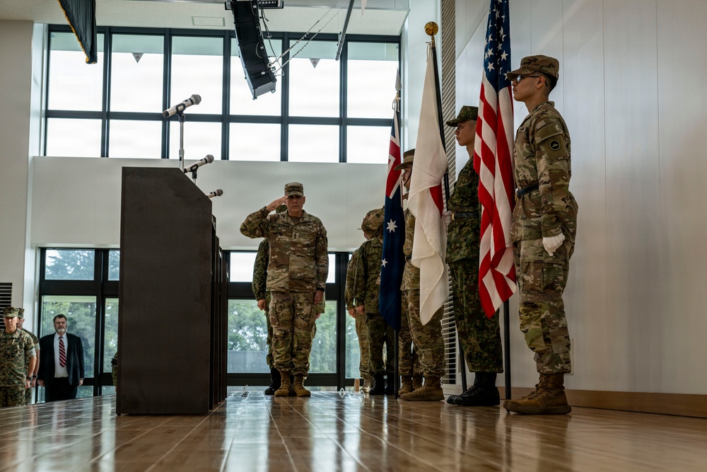 A military ceremony is taking place indoors, likely a formal event such as a change of command or allied nations gathering. A soldier in multicam uniform is rendering a salute near a podium, while a color guard holds multiple national flags including the United States, Australian, and Japanese flags. Civilian and military personnel are visible in the background observing the proceedings.
