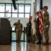 A military ceremony is taking place indoors, likely a formal event such as a change of command or allied nations gathering. A soldier in multicam uniform is rendering a salute near a podium, while a color guard holds multiple national flags including the United States, Australian, and Japanese flags. Civilian and military personnel are visible in the background observing the proceedings.