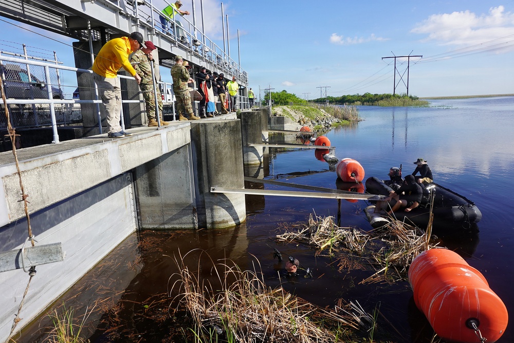 The image shows a water control structure or flood gate where a group of personnel, including military and civilian workers, are observing divers in the water below. Divers in black wetsuits are working near the base of the concrete structure, accompanied by large orange buoys and what appears to be a metal boom or barrier. The scene suggests an underwater inspection, maintenance, or debris-clearing operation at a water management facility.