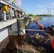 The image shows a water control structure or flood gate where a group of personnel, including military and civilian workers, are observing divers in the water below. Divers in black wetsuits are working near the base of the concrete structure, accompanied by large orange buoys and what appears to be a metal boom or barrier. The scene suggests an underwater inspection, maintenance, or debris-clearing operation at a water management facility.