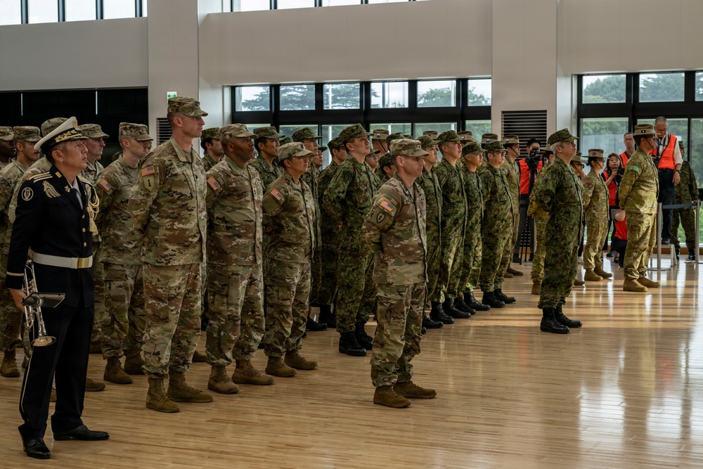 A formal military ceremony or formation is taking place inside a large indoor facility with hardwood floors. Multiple military personnel from what appear to be different nations are standing at attention in organized rows, suggesting a joint military exercise or official ceremony. A musician in dress uniform is visible on the left side, indicating a ceremonial event.