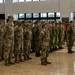 A formal military ceremony or formation is taking place inside a large indoor facility with hardwood floors. Multiple military personnel from what appear to be different nations are standing at attention in organized rows, suggesting a joint military exercise or official ceremony. A musician in dress uniform is visible on the left side, indicating a ceremonial event.