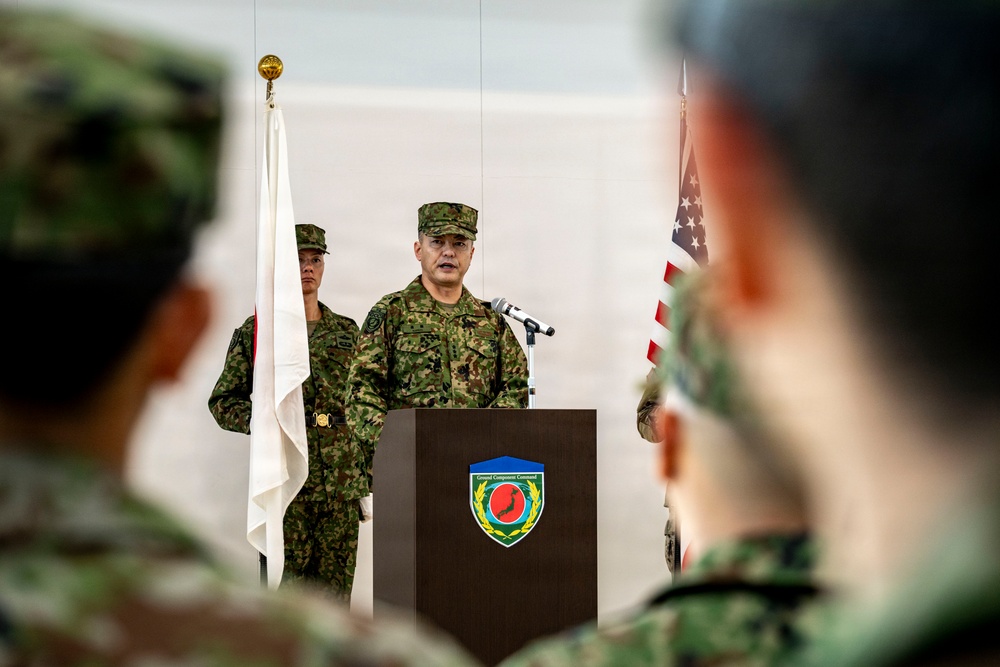 A military officer in Japanese Ground Self-Defense Force camouflage uniform is delivering a speech at a podium during a formal ceremony. The podium bears a shield-shaped insignia that reads 'Ground Component Command.' Both Japanese and American flags are visible in the background, indicating a joint military event.