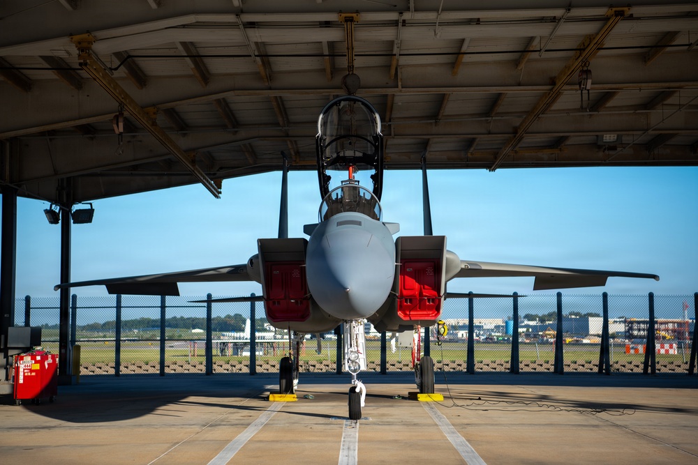 A military fighter jet, consistent with an F-15 Eagle, is parked inside an open-front maintenance hangar viewed from a direct frontal perspective. The aircraft has multiple access panels open, revealing red interior components, suggesting active maintenance or inspection work. The setting appears to be a military airfield with clear blue skies visible in the background.
