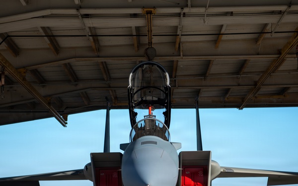 A military fighter jet, consistent with an F-15 Eagle, is parked inside an open-front maintenance hangar viewed from a direct frontal perspective. The aircraft has multiple access panels open, revealing red interior components, suggesting active maintenance or inspection work. The setting appears to be a military airfield with clear blue skies visible in the background.