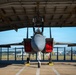 A military fighter jet, consistent with an F-15 Eagle, is parked inside an open-front maintenance hangar viewed from a direct frontal perspective. The aircraft has multiple access panels open, revealing red interior components, suggesting active maintenance or inspection work. The setting appears to be a military airfield with clear blue skies visible in the background.