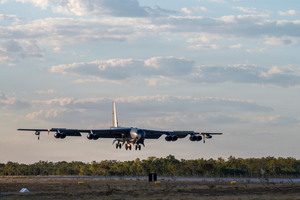 A large multi-engine strategic bomber aircraft, consistent with a B-52 Stratofortress, is captured on approach to landing with its landing gear fully extended. The aircraft is photographed head-on just above a runway, with a tree line and partly cloudy sky visible in the background. The warm lighting suggests either early morning or late afternoon conditions.
