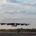 A large multi-engine strategic bomber aircraft, consistent with a B-52 Stratofortress, is captured on approach to landing with its landing gear fully extended. The aircraft is photographed head-on just above a runway, with a tree line and partly cloudy sky visible in the background. The warm lighting suggests either early morning or late afternoon conditions.