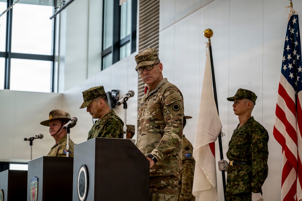 A formal military ceremony featuring personnel from multiple nations, including U.S. Army and what appears to be Japanese Ground Self-Defense Force soldiers. A senior U.S. Army officer wearing three-star rank insignia stands at a podium, suggesting a high-level command event such as a change of command or bilateral military exercise ceremony. Flags including the U.S. flag and Japanese flag are visible, held by honor guard soldiers.