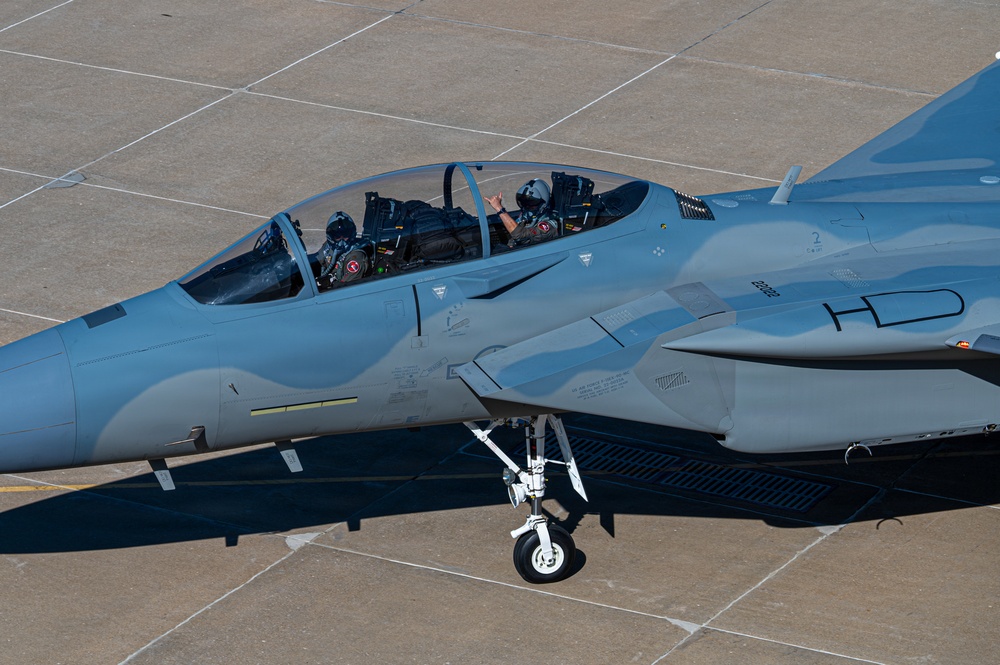 A two-seat military fighter jet, identifiable as an F-15 Eagle variant, is taxiing on a concrete airfield apron. Two crew members are visible in the tandem cockpit, with the rear occupant waving toward the camera. The aircraft bears the tail code 'HD' and is painted in an overall gray scheme.