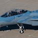 A two-seat military fighter jet, identifiable as an F-15 Eagle variant, is taxiing on a concrete airfield apron. Two crew members are visible in the tandem cockpit, with the rear occupant waving toward the camera. The aircraft bears the tail code 'HD' and is painted in an overall gray scheme.