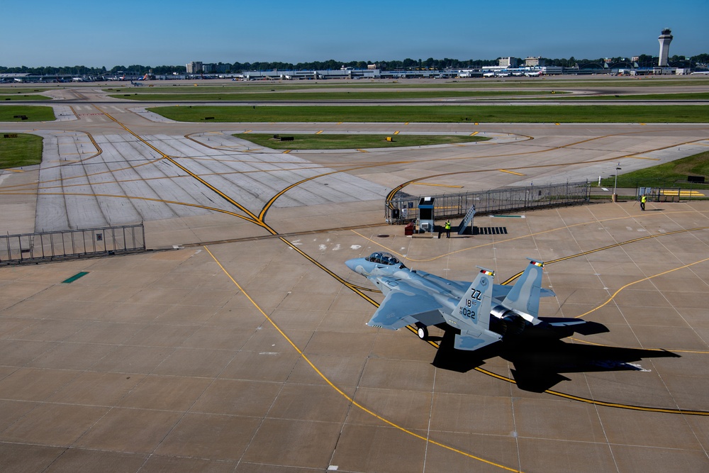 An aerial view of a military F-15 Eagle fighter jet taxiing on a large airport/air base tarmac. The aircraft bears tail code 'ZZ' and serial number '18-022', indicating it belongs to a U.S. Air Force unit. A civilian airport terminal with a control tower is visible in the background, suggesting this is a joint-use military and civilian airfield.