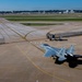 An aerial view of a military F-15 Eagle fighter jet taxiing on a large airport/air base tarmac. The aircraft bears tail code 'ZZ' and serial number '18-022', indicating it belongs to a U.S. Air Force unit. A civilian airport terminal with a control tower is visible in the background, suggesting this is a joint-use military and civilian airfield.