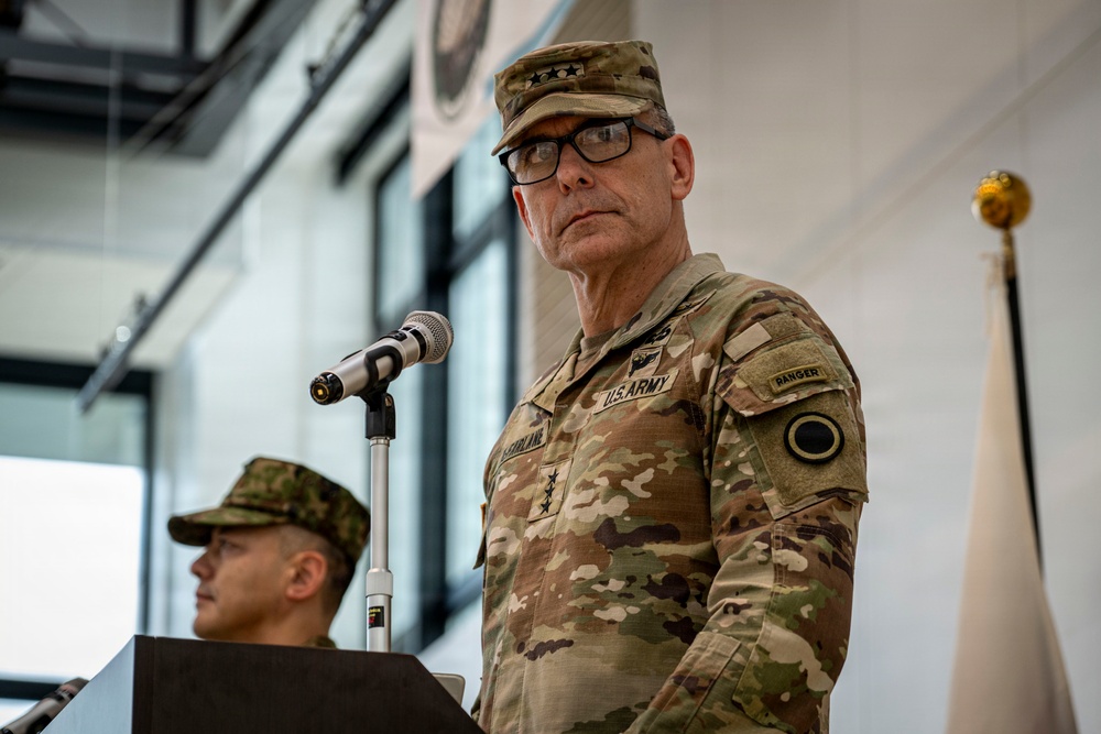 A senior U.S. Army officer wearing three-star general rank insignia stands at a podium with a microphone, appearing to deliver remarks at a formal military ceremony or event. He is dressed in OCP (Operational Camouflage Pattern) uniform with a 'RANGER' tab and U.S. Army tape visible. A second military figure in a different camouflage pattern is visible in the background.