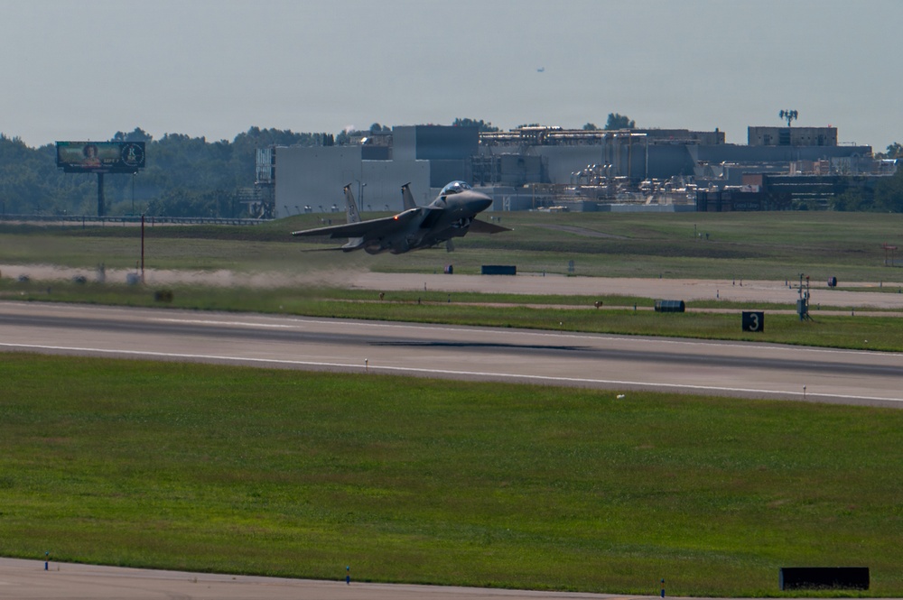 A military fighter jet, appearing to be an F-15 Eagle, is captured during takeoff or low-altitude flight over an airport runway. The aircraft is seen with afterburners engaged, generating visible exhaust and dust wake behind it. Industrial or airport facility buildings are visible in the background along with a billboard and tree line.