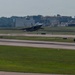 A military fighter jet, appearing to be an F-15 Eagle, is captured during takeoff or low-altitude flight over an airport runway. The aircraft is seen with afterburners engaged, generating visible exhaust and dust wake behind it. Industrial or airport facility buildings are visible in the background along with a billboard and tree line.