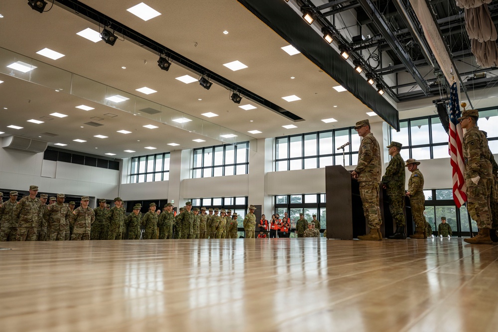 A formal military ceremony is taking place inside a large indoor gymnasium or event hall. A uniformed officer stands at a podium addressing a formation of military personnel, with an American flag displayed prominently to the right. The gathering appears to include personnel from multiple military branches or allied nations.