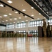A formal military ceremony is taking place inside a large indoor gymnasium or event hall. A uniformed officer stands at a podium addressing a formation of military personnel, with an American flag displayed prominently to the right. The gathering appears to include personnel from multiple military branches or allied nations.