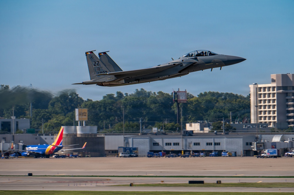 A U.S. Air Force F-15 Eagle fighter jet is captured in a low-altitude takeoff or flyby over what appears to be a joint civilian-military airfield. The aircraft displays tail codes 'ZZ' and '18', indicating it belongs to the 18th Wing based at Kadena Air Base, Japan. A Southwest Airlines Boeing 737 is visible in the background, confirming the shared civilian-military airport setting.