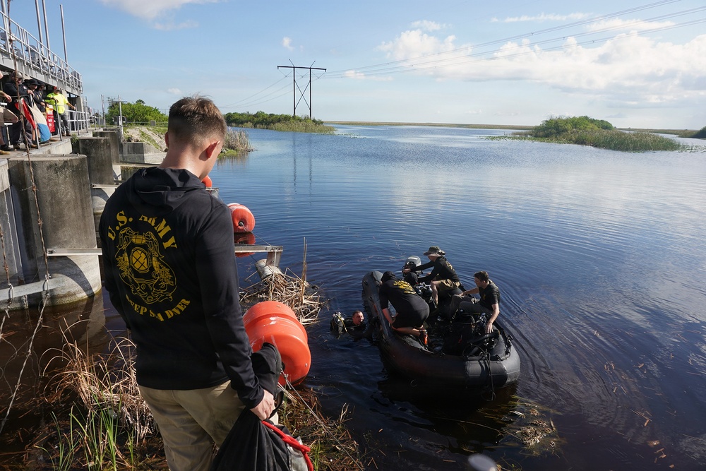 A U.S. Army dive team conducts what appears to be a water operations exercise or mission near a concrete water control structure, likely in a Florida wetlands or Everglades-type environment. Personnel in a black inflatable zodiac-style boat are assisting a diver in the water, while a soldier wearing a U.S. Army Deep Sea Diver hoodie observes from the bank holding orange dive buoys. Power transmission lines and flat wetland terrain are visible in the background.