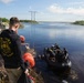 A U.S. Army dive team conducts what appears to be a water operations exercise or mission near a concrete water control structure, likely in a Florida wetlands or Everglades-type environment. Personnel in a black inflatable zodiac-style boat are assisting a diver in the water, while a soldier wearing a U.S. Army Deep Sea Diver hoodie observes from the bank holding orange dive buoys. Power transmission lines and flat wetland terrain are visible in the background.