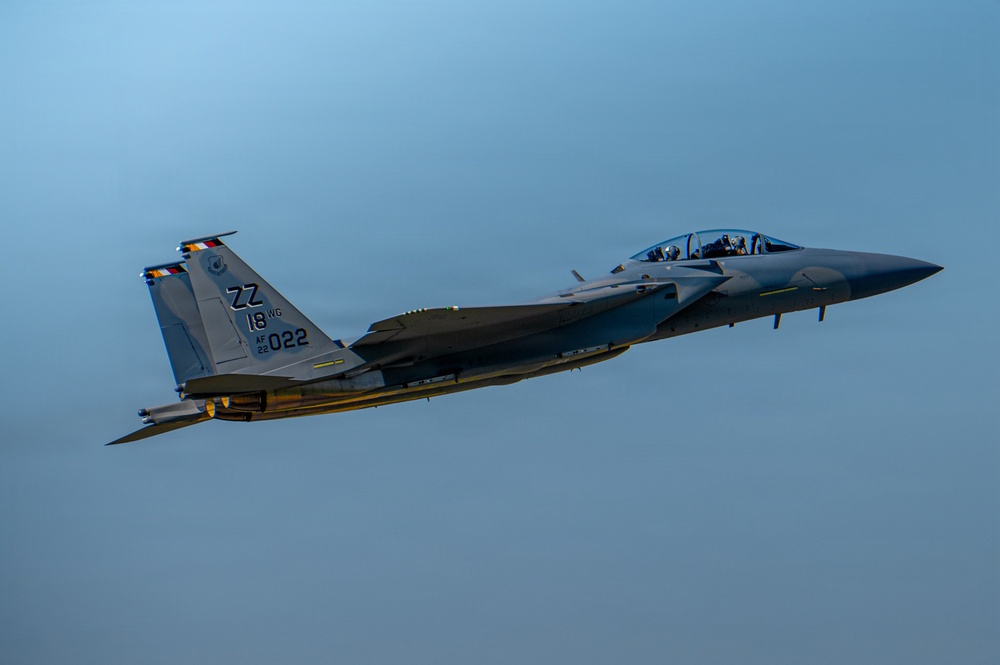 A U.S. Air Force F-15 Eagle fighter jet is captured in flight against a clear blue-gray sky. The aircraft bears tail code 'ZZ' indicating assignment to the 18th Wing at Kadena Air Base, Japan, with serial markings 'AF 22-022'. The aircraft appears to be carrying external ordnance or fuel tanks and is photographed in low-light conditions, likely at dusk or dawn.