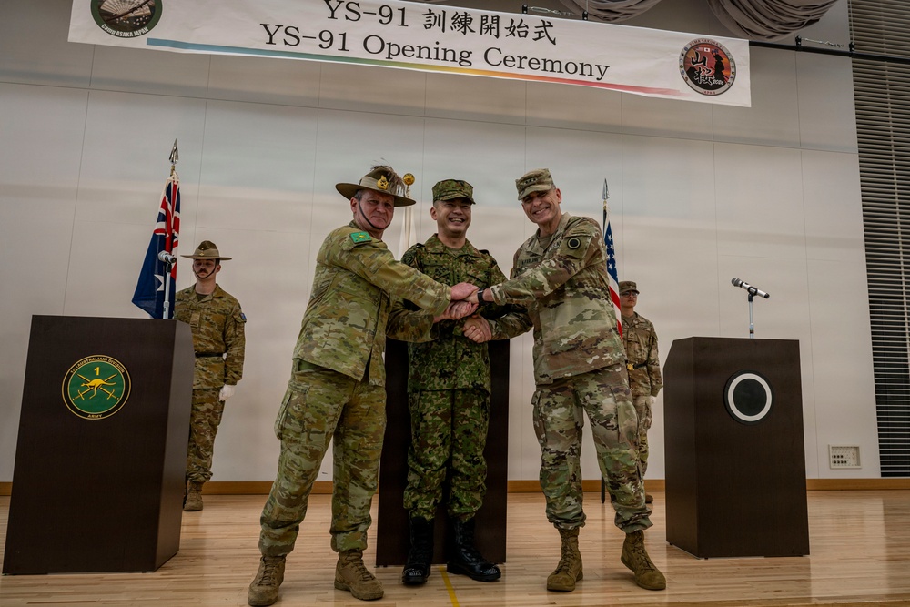 This image depicts the YS-91 Opening Ceremony, a multinational military exercise, held at Camp Asaka, Japan. Three military officers representing Australia, Japan, and the United States are shaking hands together on a stage. Flags and honor guards from the participating nations are visible in the background.