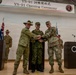 This image depicts the YS-91 Opening Ceremony, a multinational military exercise, held at Camp Asaka, Japan. Three military officers representing Australia, Japan, and the United States are shaking hands together on a stage. Flags and honor guards from the participating nations are visible in the background.