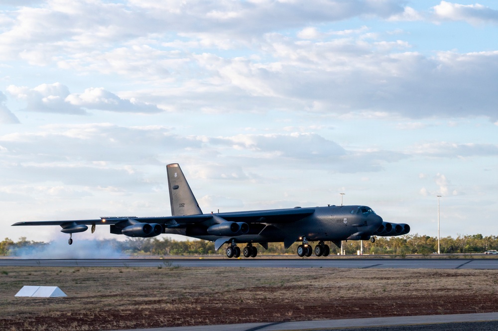 A B-52 Stratofortress strategic bomber is captured on a runway, appearing to be landing or taxiing, with visible smoke/tire smoke near the landing gear. The aircraft is painted in a dark gray scheme and displays markings consistent with the United States Air Force. The setting appears to be a civilian or joint-use airfield, likely in Australia based on the vegetation and infrastructure visible in the background.