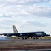 A B-52 Stratofortress strategic bomber is captured on a runway, appearing to be landing or taxiing, with visible smoke/tire smoke near the landing gear. The aircraft is painted in a dark gray scheme and displays markings consistent with the United States Air Force. The setting appears to be a civilian or joint-use airfield, likely in Australia based on the vegetation and infrastructure visible in the background.