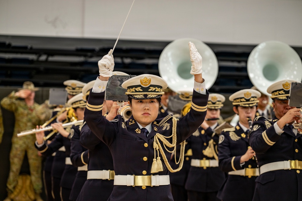 A military band performance is underway, led by a female conductor in the foreground who is actively directing with a baton and white-gloved hands raised. The band members wear dark navy dress uniforms with gold accents, white caps, and white belts, consistent with an East Asian military or ceremonial band. In the background, large sousaphones and various brass and woodwind instruments are visible, along with what appears to be a soldier in camouflage uniform observing from the left.