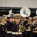 A military band performance is underway, led by a female conductor in the foreground who is actively directing with a baton and white-gloved hands raised. The band members wear dark navy dress uniforms with gold accents, white caps, and white belts, consistent with an East Asian military or ceremonial band. In the background, large sousaphones and various brass and woodwind instruments are visible, along with what appears to be a soldier in camouflage uniform observing from the left.