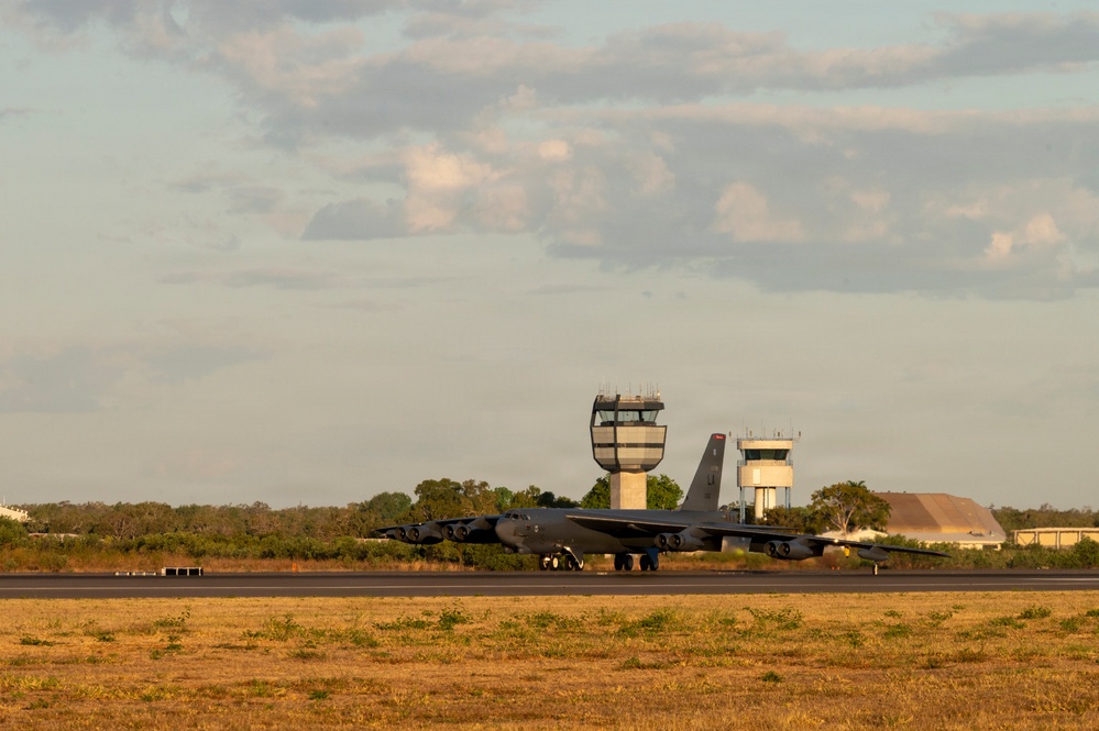 A large military bomber aircraft, identifiable as a B-52 Stratofortress, is taxiing or stationary on a runway at a military airfield. Two air traffic control towers are visible in the background, along with what appears to be a hardened aircraft shelter or hangar to the right. The image is taken during golden hour lighting conditions, suggesting early morning or late afternoon.
